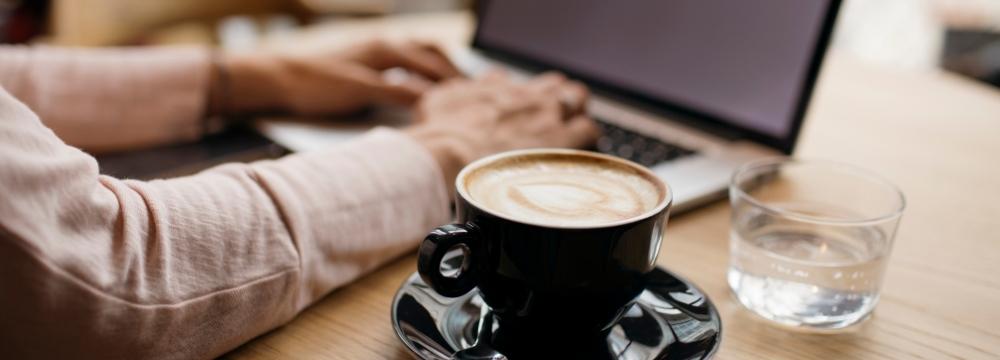 Coffee on a desk next to a woman working on laptop