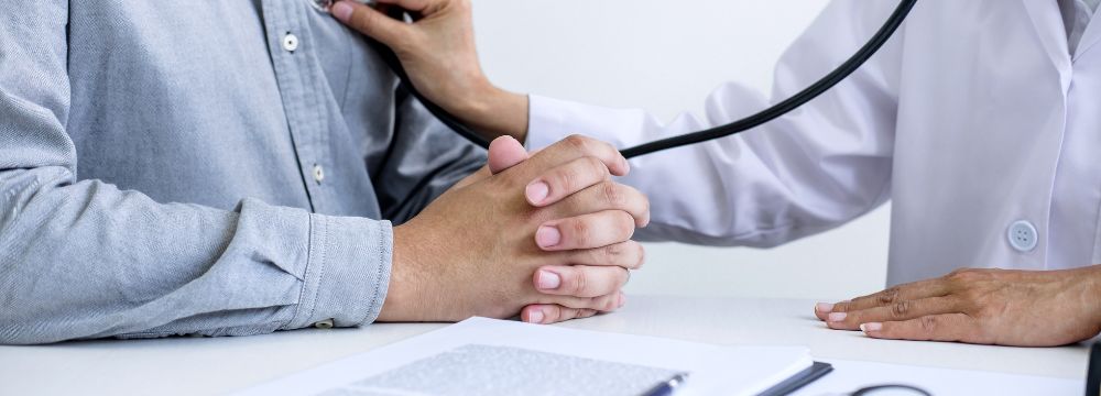 Electrophysiologist listening to male patient's heartbeat with stethoscope, paperwork on desk